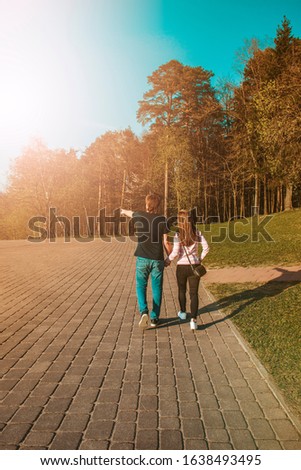 A couple in love walks hand in hand towards the sunset. Silhouette of a guy who leads a girl by the hand. Newlyweds in fashion style. Rear view. The guy points.Park with green spring grass with trees