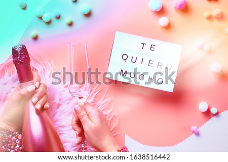 Valentine top view on pink background. Light garland, woman hands showing heart sign. Metallic pink champagne bottle. Trendy monochrome flat lay. Text "Te Quero Mucho" means I love you in Spanish.