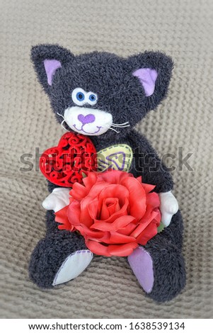 toy plush cat sits on a light background and holds a red rose and a heart.  Template for postcard to the day of lovers