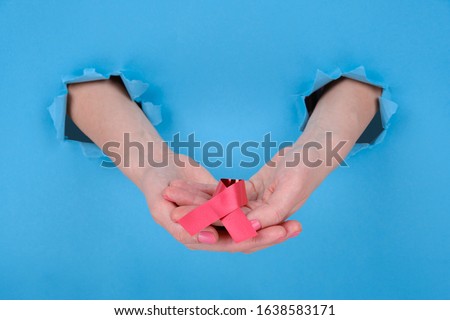 Red ribbon is symbol of fight against AIDS or disease cancer of female breast. Girl's hand holds a red ribbon on a blue torn papper background. Copy space aside for your advertising content