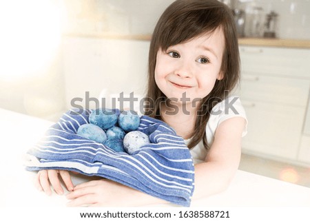 A girl sits in the kitchen and holds a basket of eggs. Blue basket with easter symbol. The child holds a lot of painted eggs. Blue eggs next to a blue towel