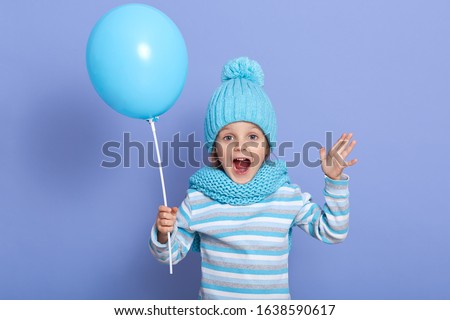 Horizontal studio picture of joyful sweet emotional little girl opening her mouth and mouth widely, raising arms, holding blue balloon, enjoying her free time, playing games. Childhood concept.