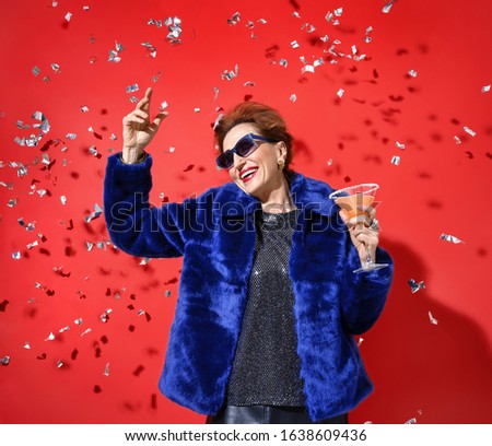 Party time. Beautiful woman dancing with glass of champagne and smiling. Photo of glamorous elderly woman in fur coat on red background