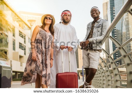 A young woman from Europe, an Arab man and an African-American man, are standing in a Dubai marina with a suitcase. Let's go travel together, say no to racism