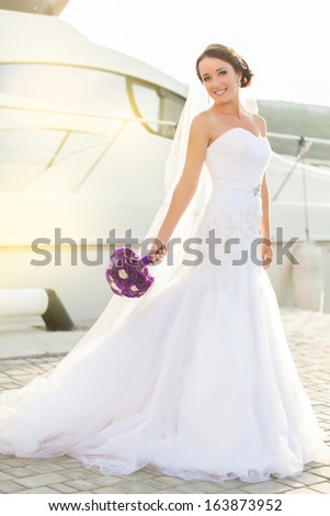 Beautiful bride in a white wedding dress poses a luxury yacht on the dock. Enjoying her wedding day.