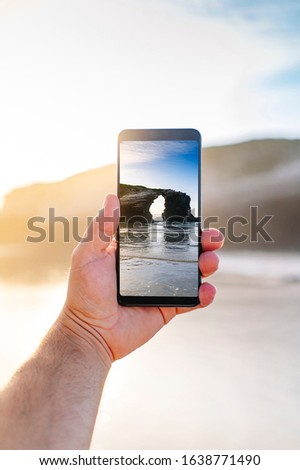 The Beach of the Cathedrals, Ribadeo Galicia. Detail of the hand of a man making a photo with a cell phone to a landscape of the beach of Las Catedrales of an arch formed by the rock in vertical
