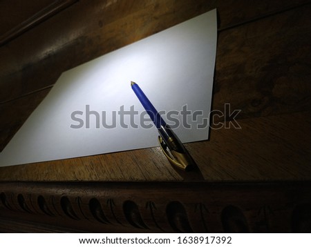 An old wooden table with decorative elements of wood on top is a white blank sheet of paper and a pen for writing and writing.