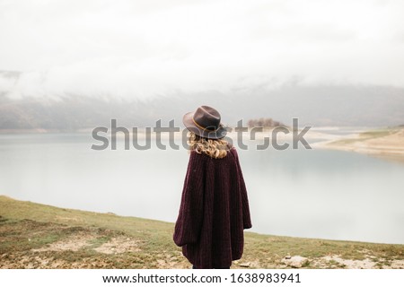 Happy beautiful woman in purple sweater holding her hat and enjoying trip on the lake. Female tourist exploring lake. She is standing turned back and looking into distance.
