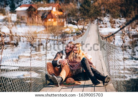Happy winter couple smiling having fun like children in winter park near a snowy river. Young beautiful woman and handsome man in winter warm casual clothes sitting back to back resting