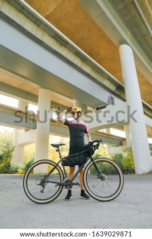 Full length portrait of a cyclist standing under a bridge with a bicycle in his hand, posing at the camera against an architecture background. Active lifestyle. Cycling is a hobby.
