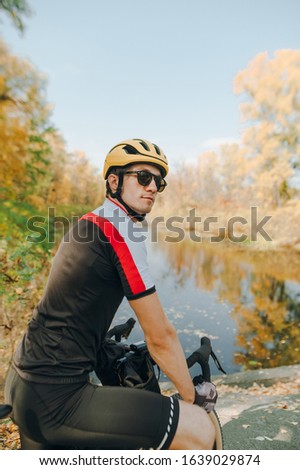Portrait of a handsome male cyclist sitting on a bike against the backdrop of a beautiful autumn landscape with a lake and looking away. Man on a bike ride in the park. Vertical photo