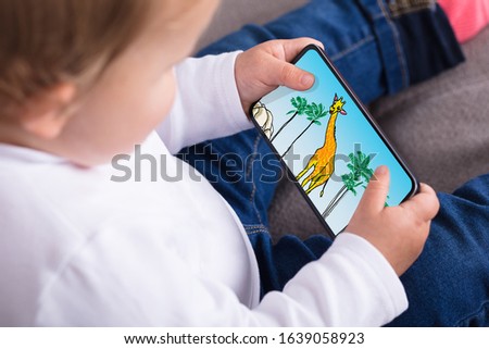 Happy Baby Girl Sitting On Sofa Watching Cartoon On Smartphone