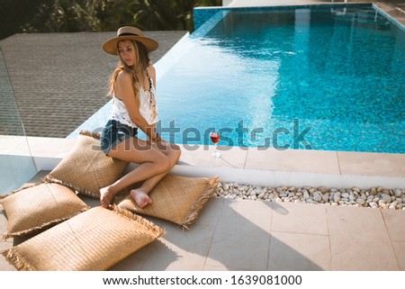 Beautiful girl in shorts and a bathing suit in a luxury villa by the pool with wine and in a hat takes cover from the sun. View of palm trees and the sea. Thailand, Samui