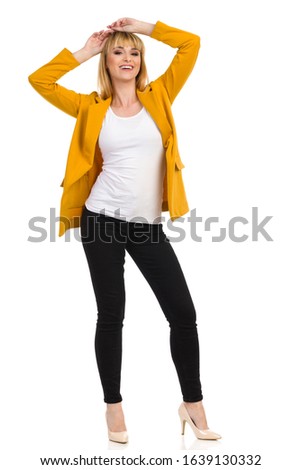 Beautiful smiling woman in unbuttoned yellow jacket is standing with arms raised and holding hands on her head. Full length studio shot isolated on white.