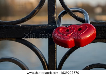 Red padlock representing composition of two hearts hanging on black ornate wrought iron railing of bridge fence on blurred background of river. Concept: Valentine's day, union of love sealed forever