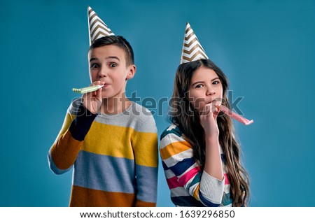 Stylish boy and cute girl with holiday caps, cones on their heads blowing whistles, celebrating a birthday isolated on a blue background.