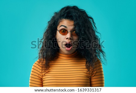 Concerned frightened curly adult woman feels fear and panic because of terrible accident, keeps mouth opened, dressed in orange turtleneck, gasps amazed, isolated on blue wall.
