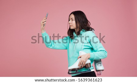 Figure skating, Hobbies and a healthy active lifestyle. A cheerful girl in a bright hoodie with skates on her shoulder is preparing to go to the skating rink. Uses a smartphone to take a selfie