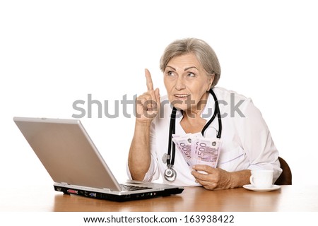 Thoughtful senior doctor sitting at table with laptop and holding euros