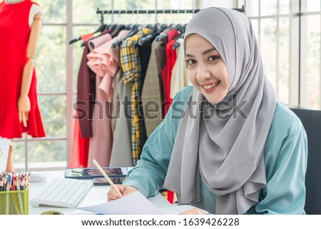 Beautiful young woman Muslim dressmaker, tailor or fashion designer in hijab is working in dressmaking studio, sewing by sewing machine looking at camera and smiling. Startup fashion business