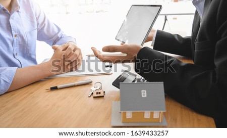 Side view of real estate agent holding  blank screen tablet presenting to customer with house model and calculator on wooden desk 