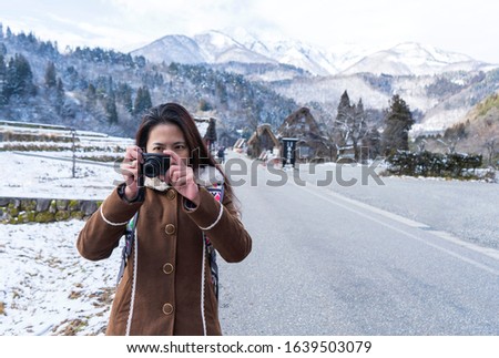 young Asian woman model with Compact Cameras wearing brown coat, black hair outdoors view of village in mountains background, winter with copy space. shirakawa go, JAPAN. Model Thai female 30s