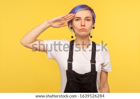 Yes sir! Portrait of serious obedient hipster girl with violet short hair in denim overalls keeping hand near temple and saluting as soldier, listening to commander. yellow background, studio shot