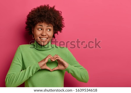 Portrait of good looking cheerful woman sends love, makes heart gesture, looks right with toothy smile, stands against pink wall with copy space for your advertising content, has romantic mood