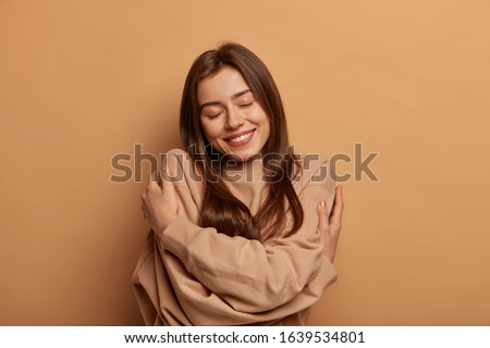 I love myself. Pretty smiling woman embraces herself gently, expresses self love, keeps eyes closed with pleasure, feels comfortable and fullfilled, being egoistic person, isolated on brown background Royalty-Free Stock Photo #1639534801