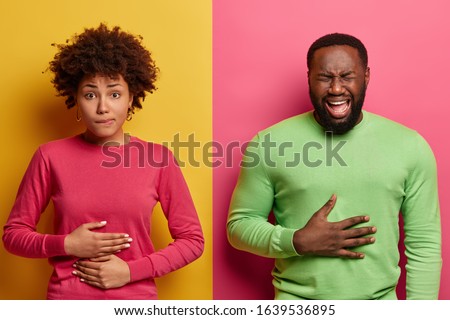 Horizontal shot of dark skinned couple touch stomachs, suffer from chronic gastritis, being hungry has displeased face expressions, isolated over yellow and pink background. Healthcare concept Royalty-Free Stock Photo #1639536895