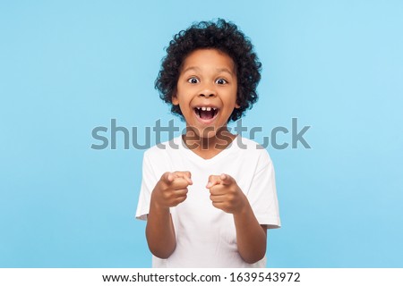 Hey you! Portrait of happy little boy with curly hair pointing finger to camera and laughing loudly with surprised face, teasing making fun of you. indoor studio shot isolated on blue background