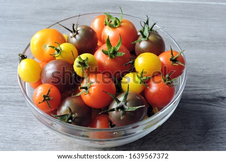 A glass bowl of ripe yellow, red and purple cherry tomatoes covered with raindrops, grey wooden background