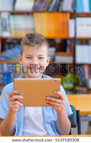 Cute boy in the library sitting with a tablet computer in his hands. Smiling while looking into the frame.