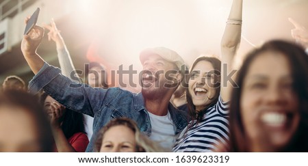 Excited couple cheering  and taking selfie while at stadium. Cheerful soccer fans taking selfie while watching match in stadium.
