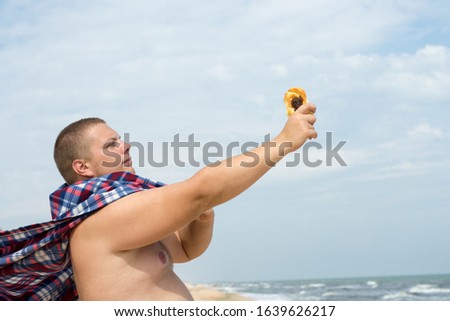 A funny fat man superhero with a plaid instead of a cloak with a bun in hand stands on the sea beach.