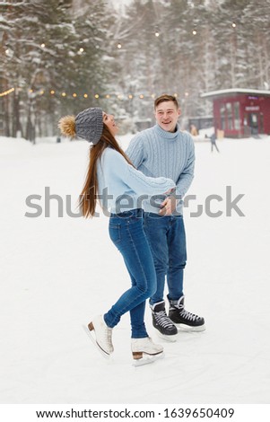 Young couple laughing ice skating on an ice rink in a winter park. Snowdrifts, trees, blurred background.