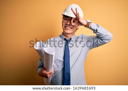 Young handsome chinese architect man wearing safety helmet holding blueprints with happy face smiling doing ok sign with hand on eye looking through fingers