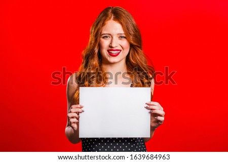 Pretty woman holding white a4 paper poster. Copy space. Smiling trendy girl with red hair on studio background.
