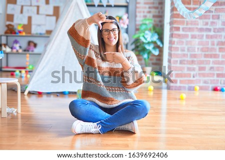 Young beautiful teacher woman wearing sweater and glasses sitting on the floor at kindergarten smiling making frame with hands and fingers with happy face. Creativity and photography concept.