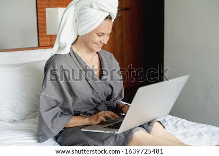 Beautiful happy young woman in a gray bathrobe and with a towel on her head on a bed in a hotel room working on a laptop, vacation business trip distant work digital nomad concept