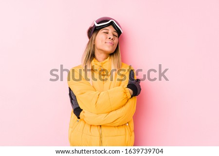 Young caucasian woman wearing a ski clothes in a pink background hugs, smiling carefree and happy.