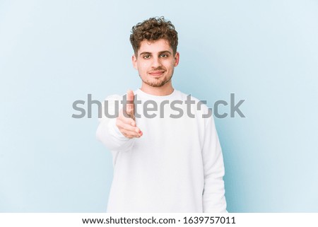 Young blond curly hair caucasian man isolated stretching hand at camera in greeting gesture.