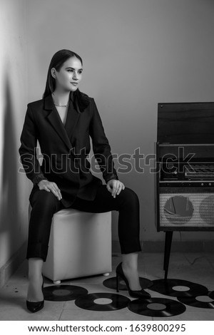 a beautiful Caucasian young woman in a black pantsuit and black sandals poses next to a vintage record player with a gramophone record in her hands. black and white picture