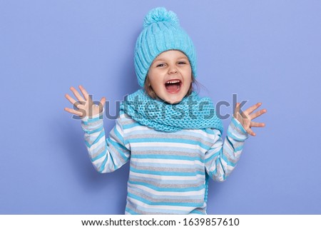 Image of cheerful positive little kid opening eyes and mouth widely, raising arms, being playful, wearing blue hat and scarf, striped sweatshirt, standing isolated over lilac background in studio.