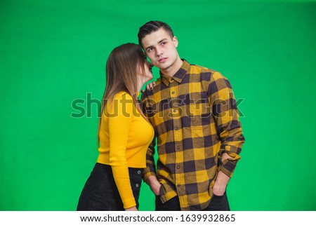 Young women sharing gossip and whispering secrets to her boyfriend, he is looking with puzzled face expression, standing over green background.