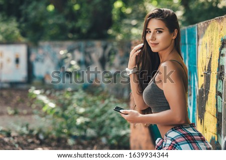 Beautiful smiling girl hipster posing using mobile phone near the wall with graffiti