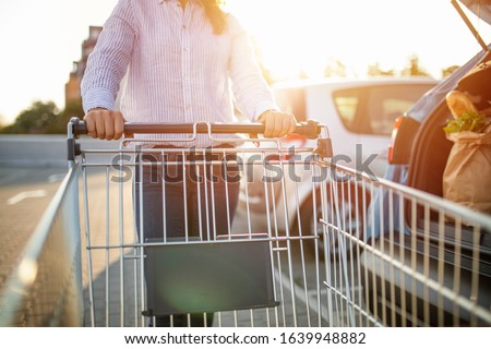 Woman with bags and shopping cart loads a car. Woman putting bags into car after shopping. Woman pushing supermarket cart in urban public parking