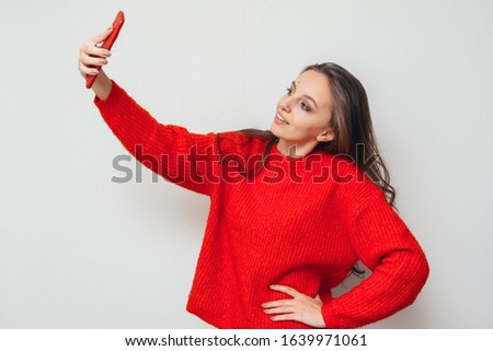 Young girl in a red sweater makes selfie on the phone against the background of a white wall.
