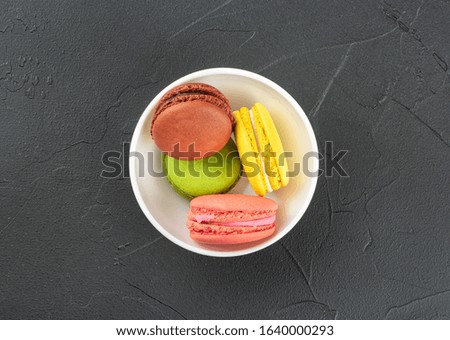 Multicolored macaroons in a white bowl on a dark background, top view