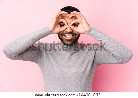 Young latin man against a pink background isolated showing okay sign over eyes